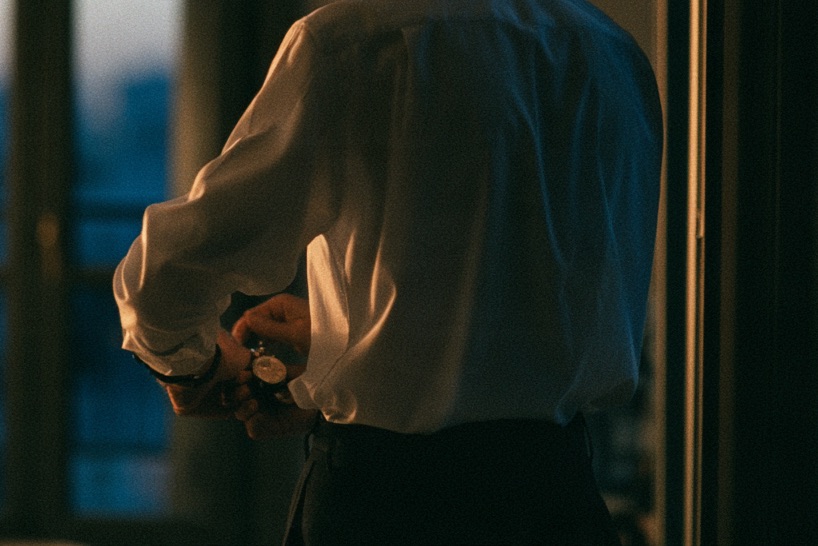 A man in a white shirt puts on his watch by the window, at dawn.