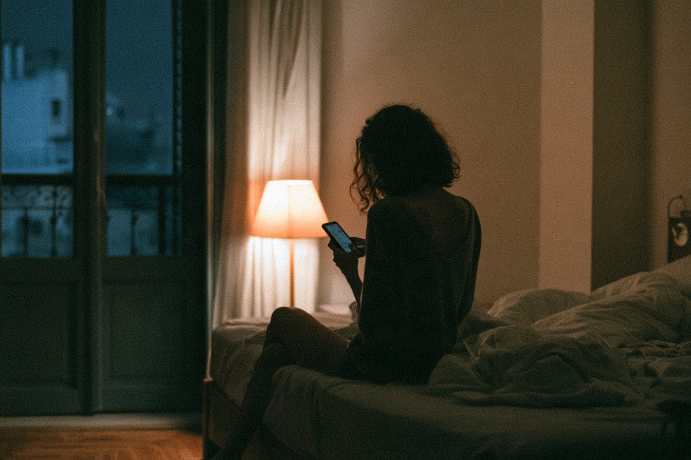 A woman sits on the edge of the bed at night, looking at her phone.